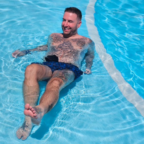 Nathan lounging in a swimming pool and smiling wearing a black stoma bag and blue swimming trunks