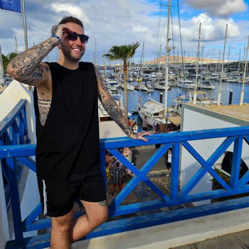 Nathan leaning on a railing at a marina in a tropical country wearing shades, a black vest and black shorts and looking relaxed